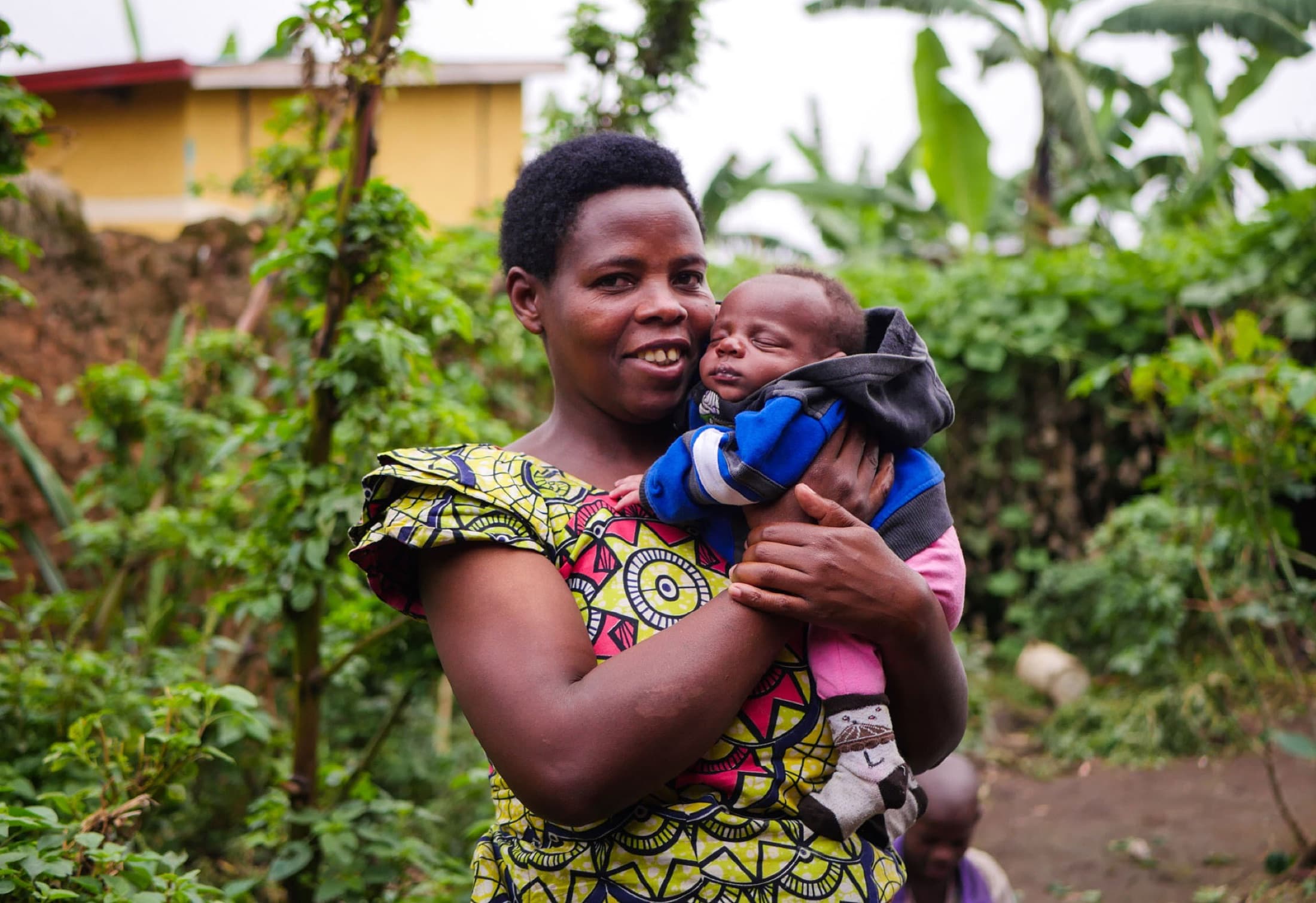 A laughing mother in bright kitenge holds her newborn outside the Atiak birth center